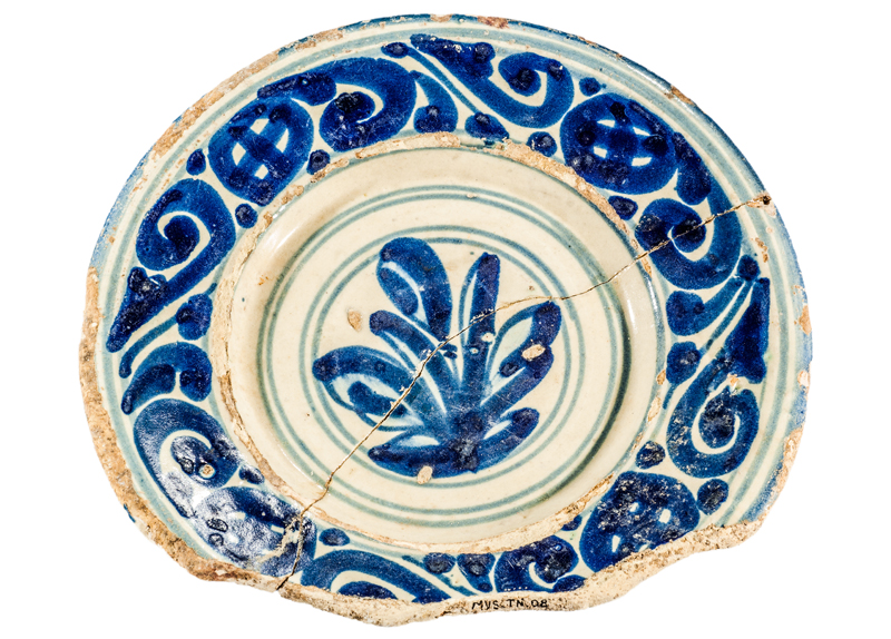 Plat de ceràmica vidrada decorada en blau. Producció catalana. Presenta una decoració en orla a la vora a base decoració de branquillons de volutes i roda de carreta. A la part central, decoració vegetal. segle <span style=\