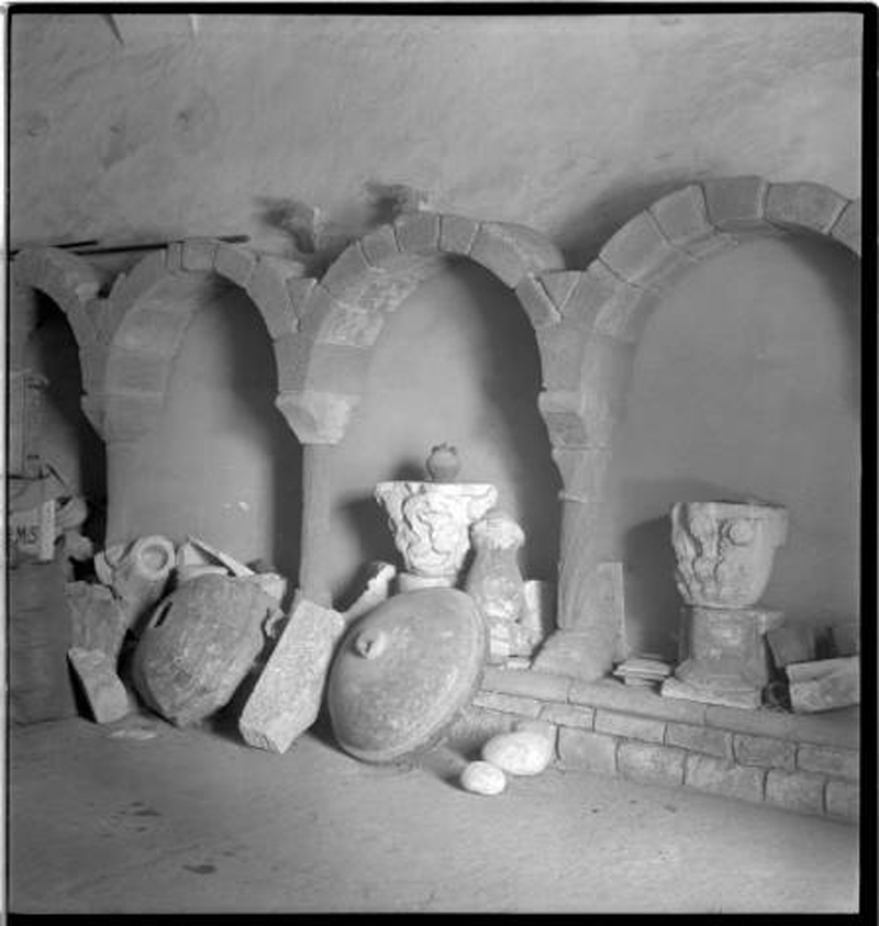 Parts de la galeria romànica del claustre i altres elements al magatzem on foren guardats després de la seva localització durant  els treballs de desenrunament de l’església. Foto: Família Cuyàs. Institut Cartogràfic de Catalunya (sense data). 